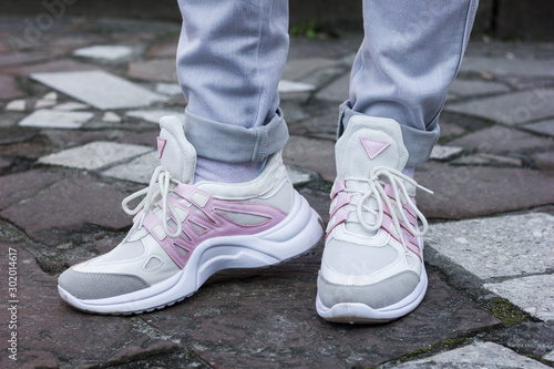 beautiful white sneakers with gray and pink closeup on a female leg in gray jeans on a background of street tile