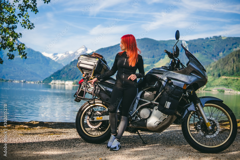 Obraz premium Pretty women is standing with traveler motorcycle. Vacation and hobby concept, jorney on two wheels. Sunny summer day in the Alpine mountains. Zell am see lake on background Austria. Copy space