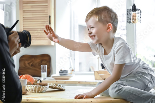 Portrait of aborable boy model posing in front of photographer. Kid sitting on table in bright kitchen room. photo