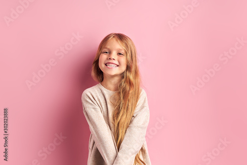 Portrait of positive cheerful girl cutely smiling at camera, girl with long golden hair in white blouse. Pink background