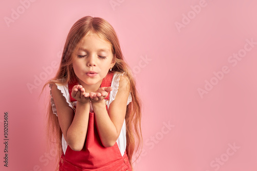 Portrait of little caucasian girl with long hair and closed eyes sending, blowing kiss to camera, wearing red overalls isolated over pink background.