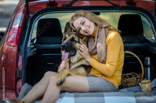 A happy Caucasian girl sits in trunk of a car, hugs a German shepherd dog and smiles, a cheerful playful teen puppy looks at the camera. Friendship of man and animal, travel, camping. Autumn forest