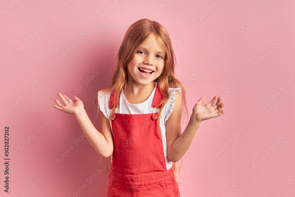 Funny laughing caucasian girl dressed in red overalls stand looking at camera on pink background Funny laughing caucasian girl dressed in red overalls stand looking at camera on pink background