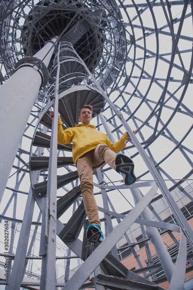 A young guy in a yellow hoodie and turquoise sneakers stands on a spiral staircase. Teen jumps off with a beautiful spiral staircase in the beautiful architecture