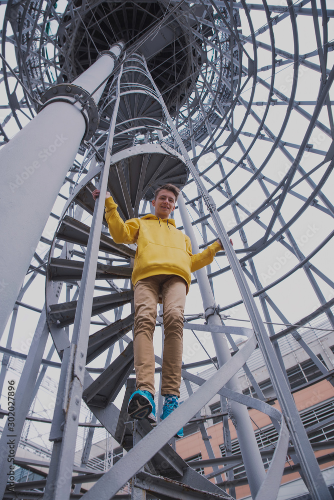 A young guy in a yellow hoodie and turquoise sneakers stands on a spiral staircase. Teen jumps off with a beautiful spiral staircase in the beautiful architecture A young guy in a yellow hoodie and turquoise sneakers stands on a spiral staircase. Teen jumps off with a beautiful spiral staircase in the beautiful architecture