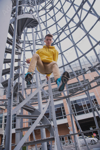 A young guy in a yellow hoodie and turquoise sneakers stands on a spiral staircase. Teen jumps off with a beautiful spiral staircase in the beautiful architecture © Антон Фрунзе