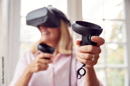 Mature Disabled Woman In Wheelchair At Home Wearing Virtual Reality Headset With Gaming Controllers