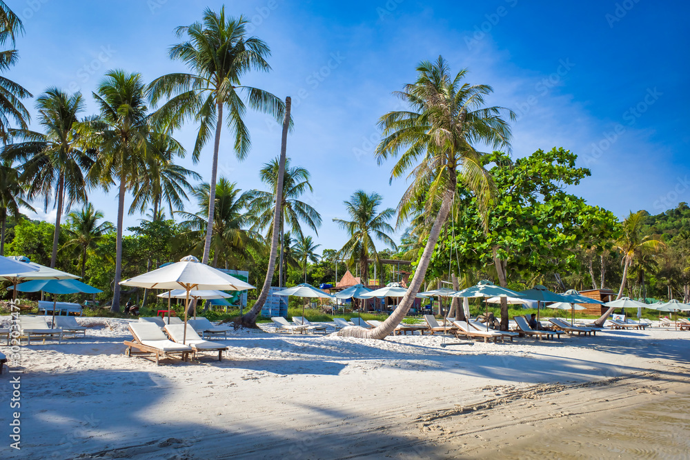 Sandy beach on the bay, high palm trees, blue sky, sun loungers for relaxation and sunbathing under umbrellas, tourists resting, Phu Quoc island, Vietnam