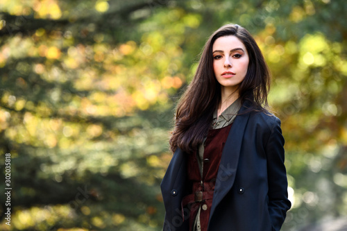 Autumn portrait of an attractive young woman in elegant coat standing in the park with beautiful foliage on the background.