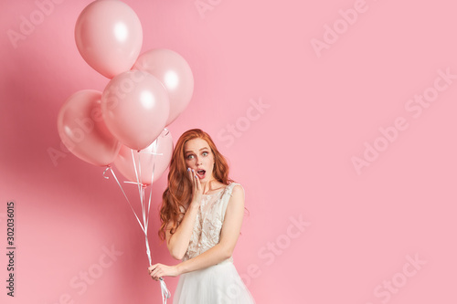 Portrait of surprised young woman dressed in white dress, auburn hair holding pink air balloons isolated over pink background