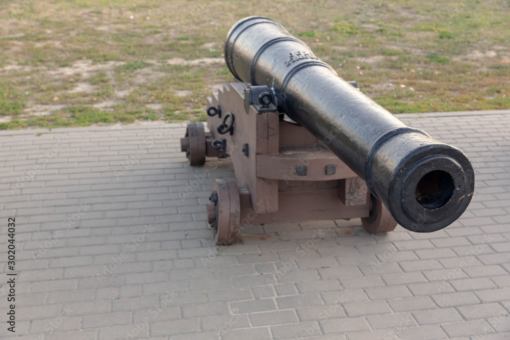 An ancient, black, cast-iron gun on a wooden, brown, painted carriage stands on the gray-tiled pavement. An ancient, black, cast-iron gun on a wooden, brown, painted carriage stands on the gray-tiled pavement.