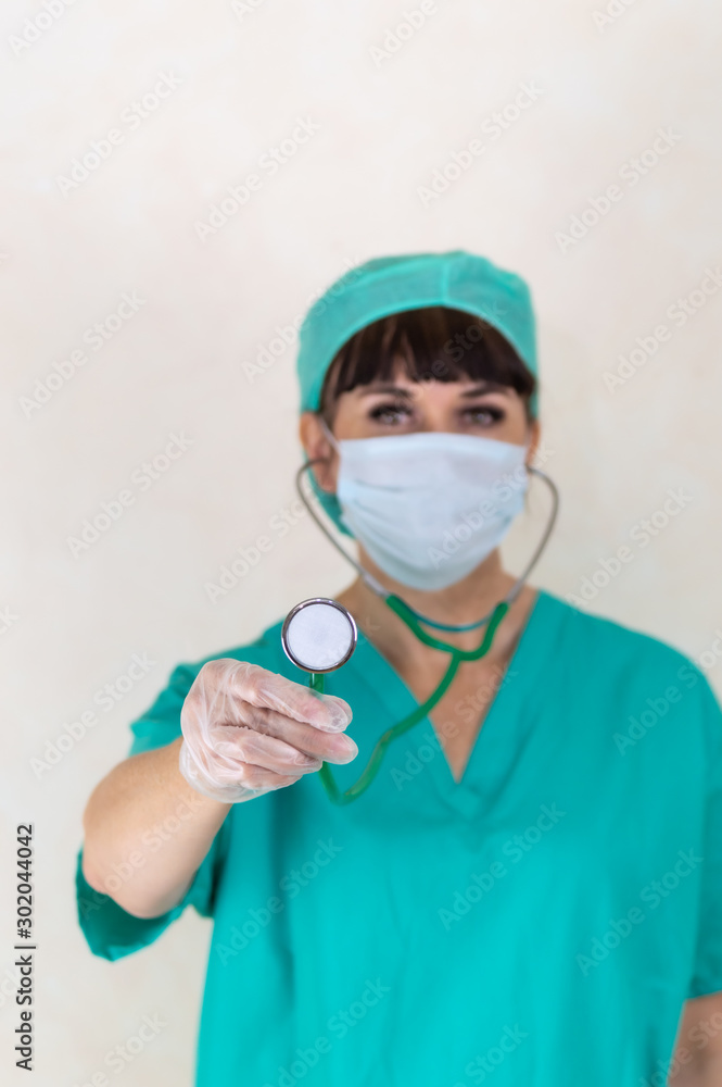 Blurred female doctor or nurse wearing face mask, hair net and green scrubs. Focus on stethoscope and hand with latex glove. Making eye contact. Plain pale background with copy space. Blurred female doctor or nurse wearing face mask, hair net and green scrubs. Focus on stethoscope and hand with latex glove. Making eye contact. Plain pale background with copy space.