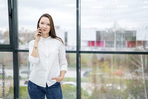 A woman in a white shirt stands by the window and talks on the phone. European girl is calling on a smartphone and smiling.