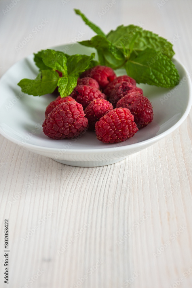 Raspberries with mint leaves on white plate and white wood in vertical with copy space Raspberries with mint leaves on white plate and white wood in vertical with copy space