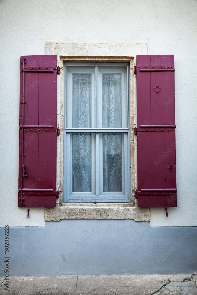 Blue window with crimson coloured traditional wooden shutters and lace curtains of a house on a street in Arles, south of France.