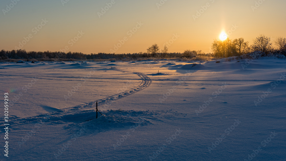 Setting sun by the winter sea shore with tracks and fish traps in the snow