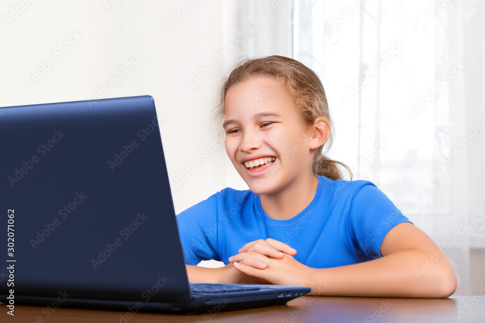 Beautiful cute little girl sitting at a computer and having fun laughing while looking at a laptop monitor. Distance learning, chatting with friends, enterta Beautiful cute little girl sitting at a computer and having fun laughing while looking at a laptop monitor. Distance learning, chatting with friends, enterta