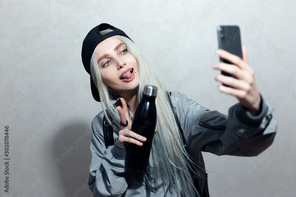 Portrait of young blonde girl with thermo bottle in hand take a selfie photo on smartphone.