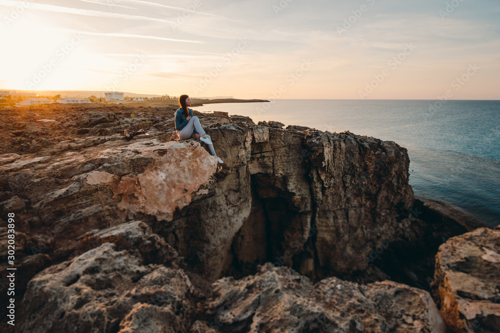 Woman traveler sitting near sea on top of cliff in summer mountains and enjoying view of sea and nature. Cape Greco, Cyprus, Mediterranean Sea. Sunrise Woman traveler sitting near sea on top of cliff in summer mountains and enjoying view of sea and nature. Cape Greco, Cyprus, Mediterranean Sea. Sunrise