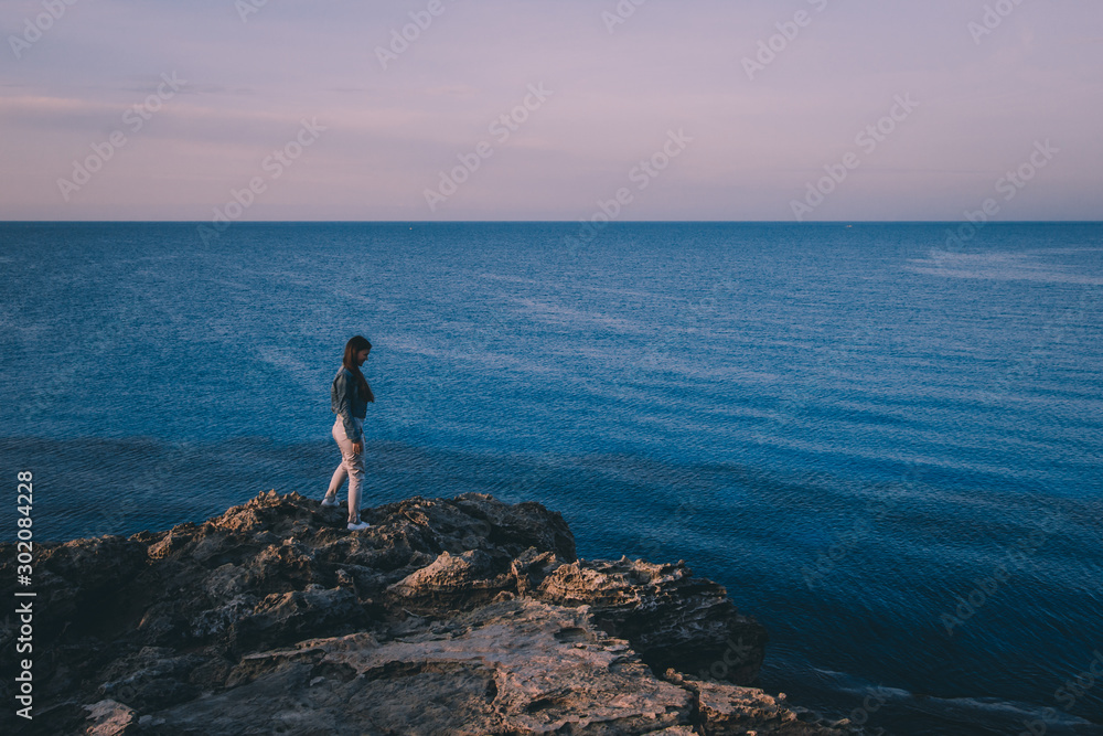 Woman traveler standing near sea on top of cliff in summer mountains and enjoying view of sea and nature. Cape Greco, Cyprus, Mediterranean Sea. Sunrise Woman traveler standing near sea on top of cliff in summer mountains and enjoying view of sea and nature. Cape Greco, Cyprus, Mediterranean Sea. Sunrise