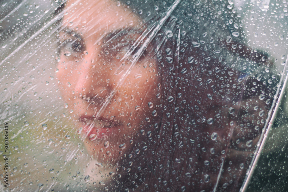 Blurred sad face of a girl behind transparent umbrella with raindrops. Close-up