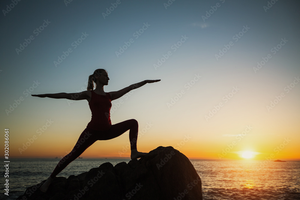 Silhouette yoga woman doing fitness exercises near the sea at sunset.
