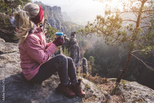 Female blond hiker outdoor clothing with coffee cup in hands on limestone rock enjoying sunset back lit view of mountain ridge and forest down the valley. Travel lifestyle adventure concept