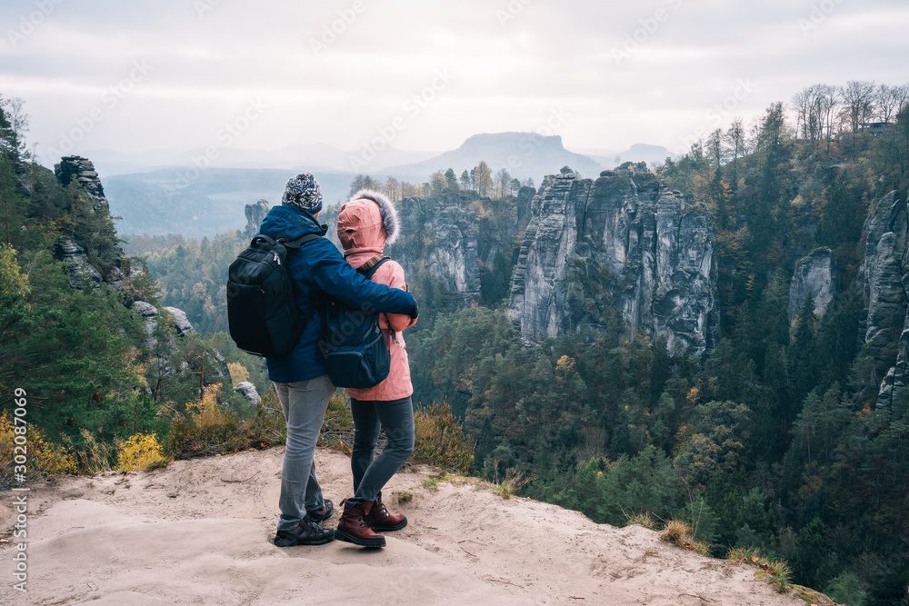 Young couple in outdoor clothing with backpacks standing on plateau enjoying view of mountain ridge and wild forest in the valley on hiking trail. Travel lifestyle concept