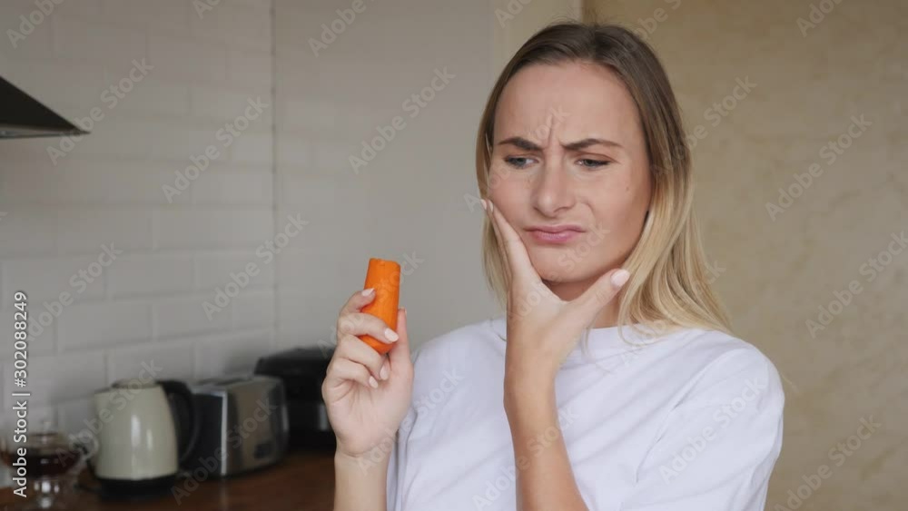 Young attractive woman eating carrots having horrible tooth ache while sitting at home in the living room Young attractive woman eating carrots having horrible tooth ache while sitting at home in the living room