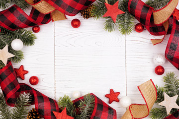 Christmas frame of ornaments, branches, burlap and red and black buffalo plaid ribbon. Overhead view on a white wood background.