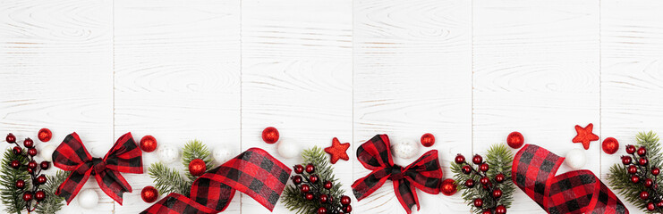 Christmas border banner of ornaments, branches and red and black checked buffalo plaid ribbon. Top view on a white wood background.