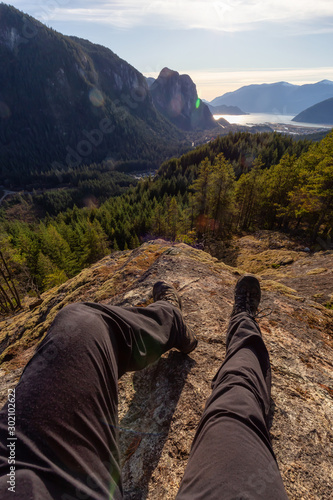 Sitting on the edge of a cliff and enjoying the Beautiful Canadian Mountain Landscape View during a sunny evening. Taken in Squamish, North of Vancouver, British Columbia, Canada.