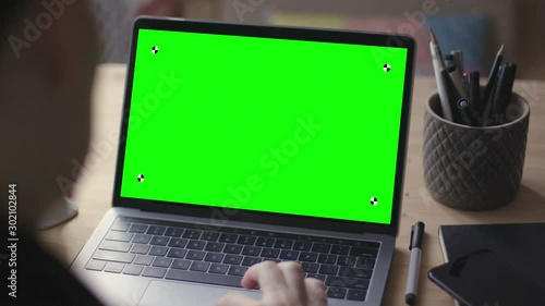 Over the shoulder shot of young woman working on modern laptop with green screen mock-up. Hands on keyboard.