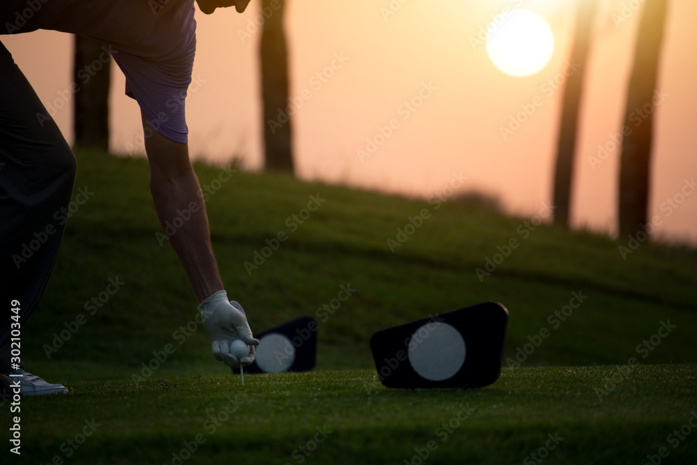 Obraz premium The golfer's hand grips the golf ball and tee presses down at starting point amid sunrise in morning.