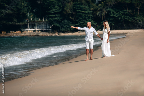 Attractive trendy young couple in white clothes are walking along on the beach and holding hands. Beautiful blonde with long hair and a bald man are on vacation. Phuket. Thailand.