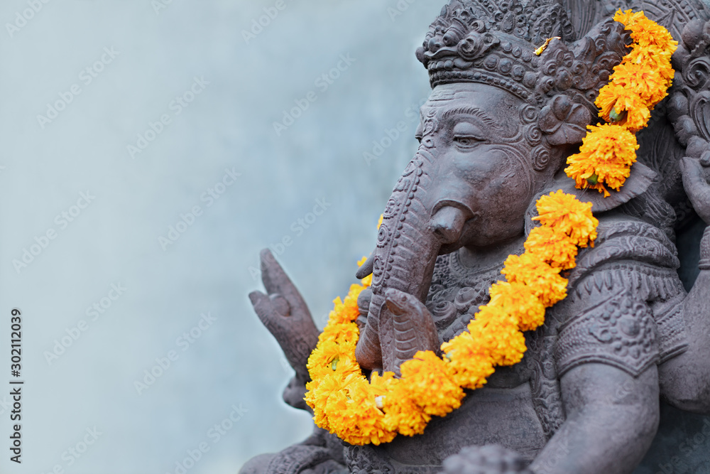 Fototapeta Ganesha sitting in meditating yoga pose in front of hindu temple. Decorated for religious festival by orange flowers garland, ceremonial offering. Balinese travel background. Bali island art, culture.