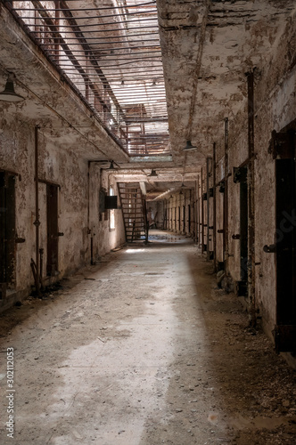 Hallway of an old abandoned prison showing doorways to cells, metal stairs, and bars on the ceiling.