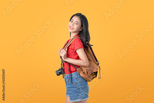 Smiling happily Asian woman traveler holding camera and bag on orange background.