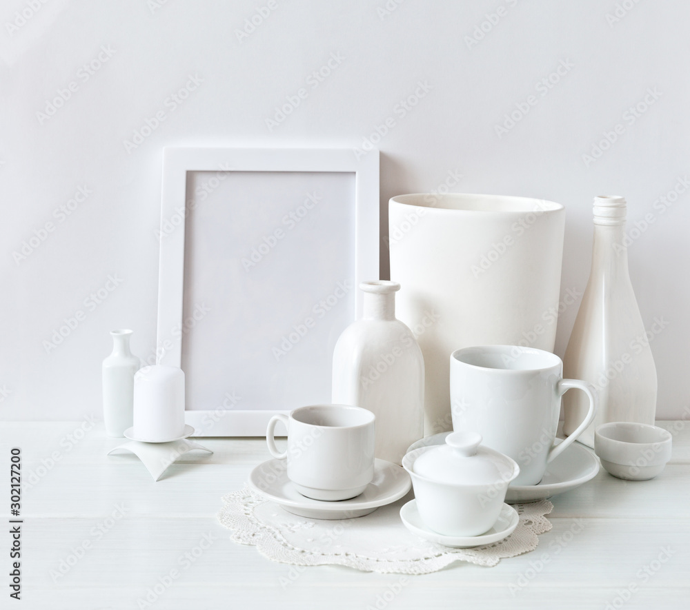 Still life of white objects: cups with saucers, vases, an empty frame and candles on a white background. Place for text, soft light, high key. Still life of white objects: cups with saucers, vases, an empty frame and candles on a white background. Place for text, soft light, high key.