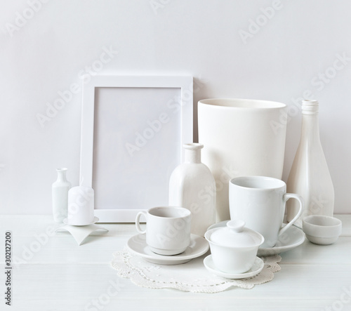 Still life of white objects: cups with saucers, vases, an empty frame and candles on a white background. Place for text, soft light, high key.