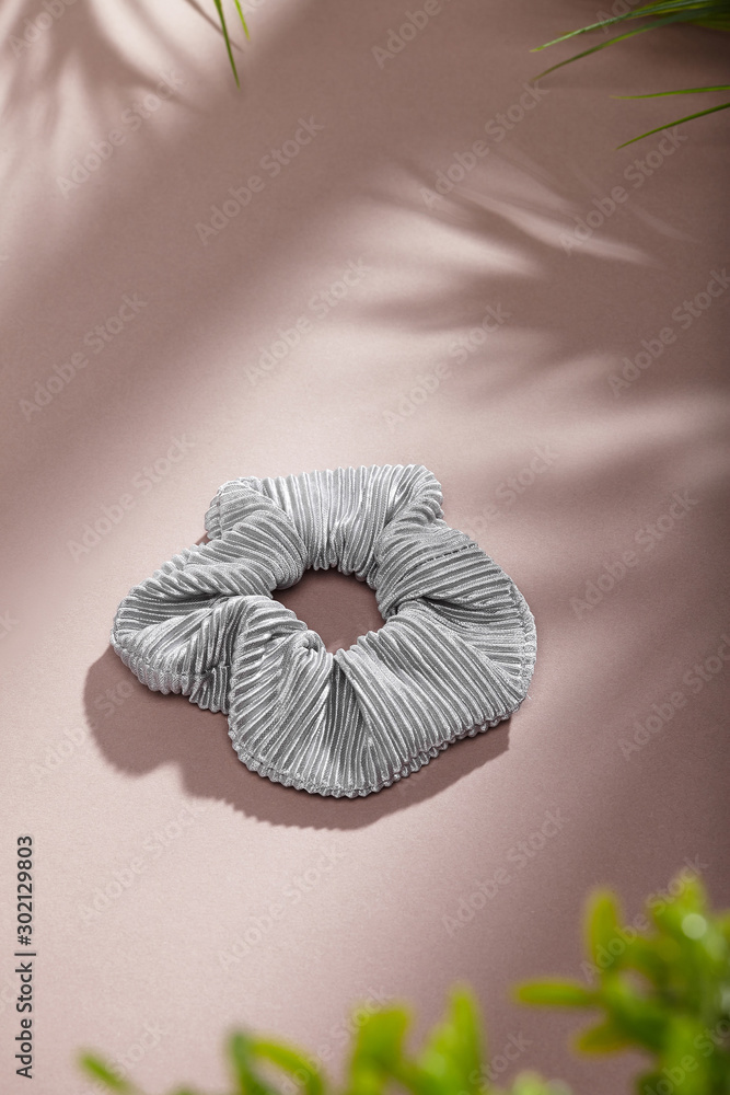 Obraz premium Object photo of a grey textured scrunchie. The scrunchie is lying on a beige background. There are leaves in corners of a photo. The scrunchie and leaves casting a shadow.