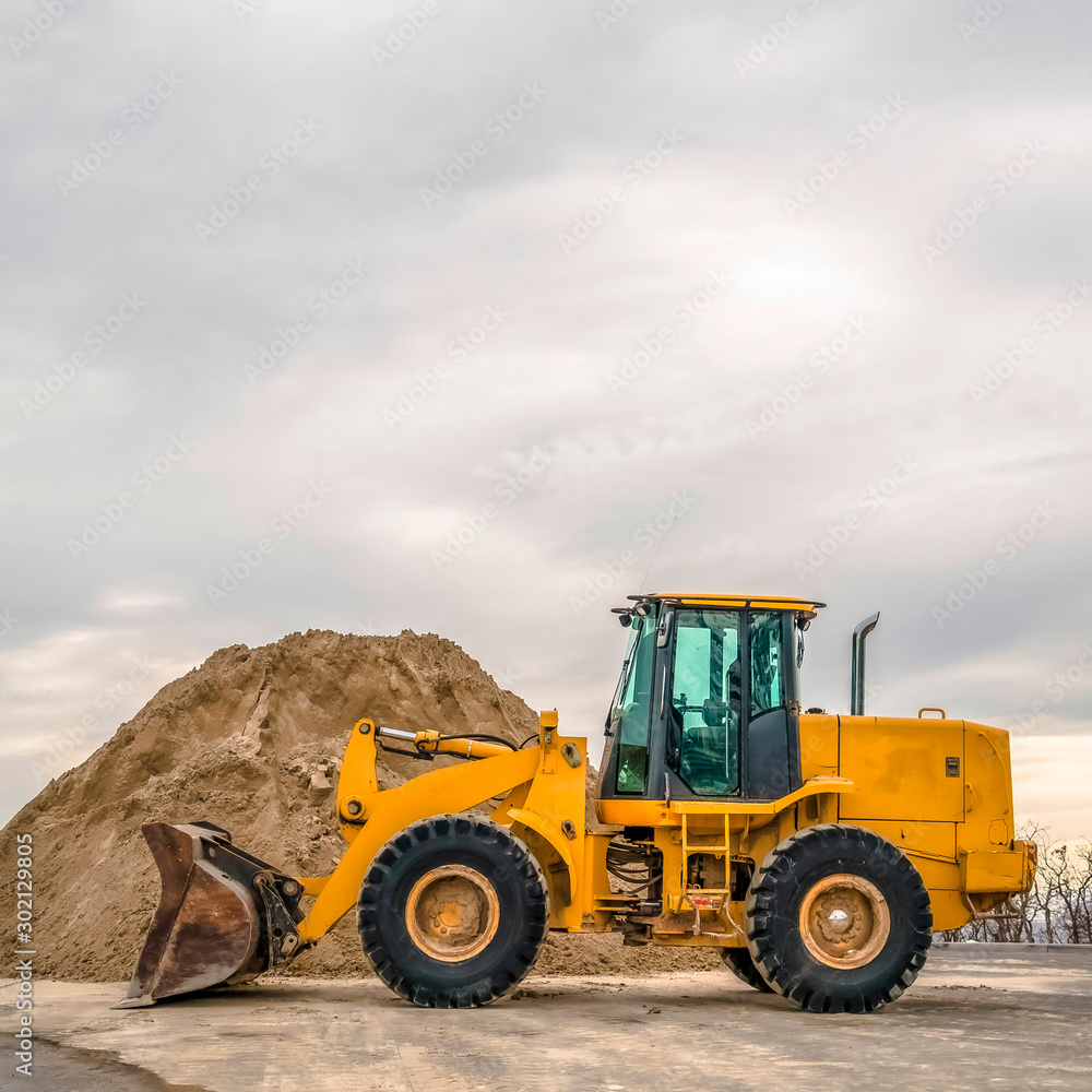 Square frame Bulldozer in front of a mound of soil with leafless trees and cloudy sky background