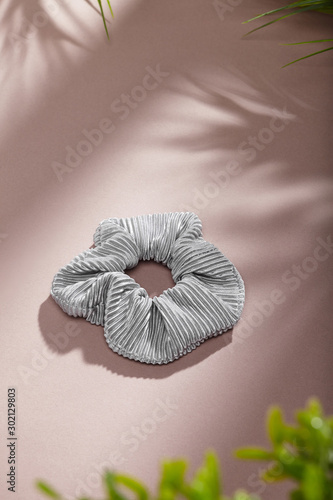 Object photo of a grey textured scrunchie. The scrunchie is lying on a beige background. There are leaves in corners of a photo. The scrunchie and leaves casting a shadow.