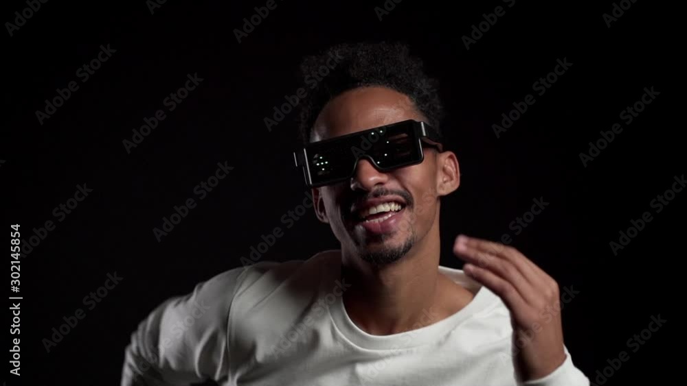 African man in glowing neon glasses moving to music rhythm on black studio background. Dancing positive comic guy. Slow motion. African man in glowing neon glasses moving to music rhythm on black studio background. Dancing positive comic guy. Slow motion.