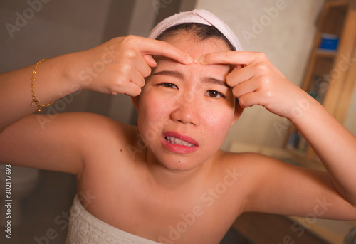 lifestyle face portrait of young upset and expressive Asian Chinese woman squeezing pimples while looking at the mirror in the bathroom in facial skin care and beauty