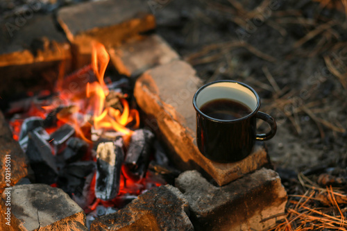Black enamel cup of hot drink sitting on a stone by the fire in the open air. Focus on a mug with a blurred background.Detail of a camping, lifestyle while traveling. Hot coffee at the campsite