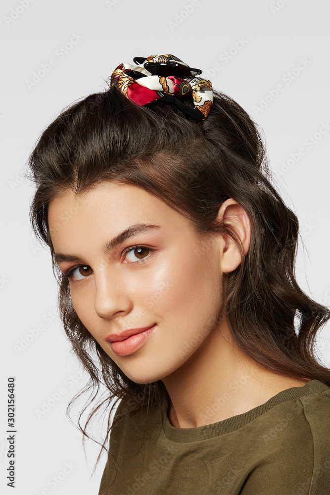 Naklejka premium Closeup half-turn face portrait of girl with dark wavy hair, wearing khaki t-shirt. Her hair is pulling with black hairpin crab, decorating with white patterned bow.