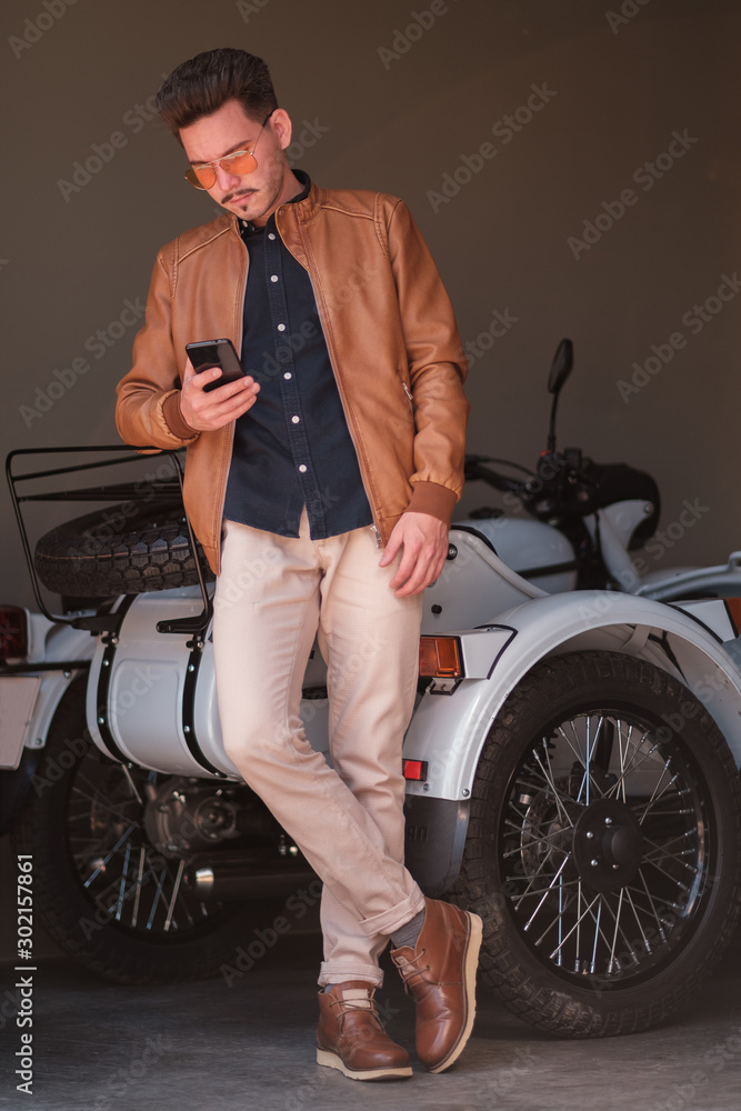 Obraz premium young guy stands with a smartphone in his hands on a background of a motorcycle. casual style. portrait of a successful man.