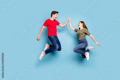 Great job. Full length profile side photo of cheerful two romantic people jump give highfive celebrate victory wear modern t-shirt denim jeans sneakers isolated blue color background