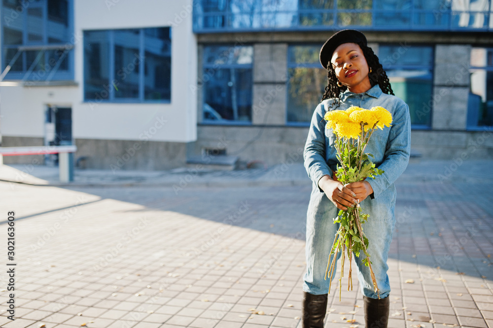 Fototapeta premium Stylish fashionable african american women in jeans wear and black beret with yellow flowers bouquet posed outdoor in sunny day against blue modern building.